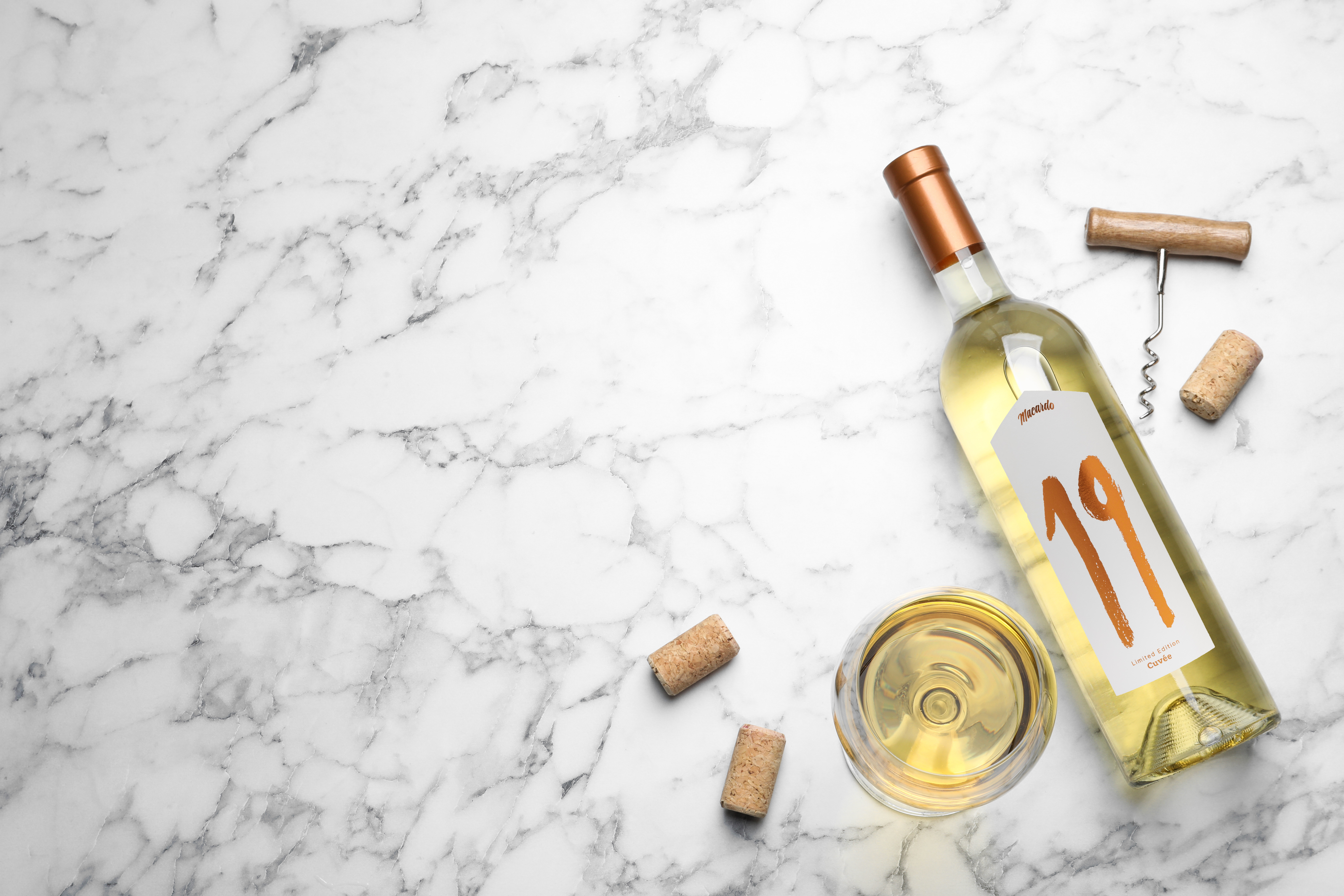 Auf einer Marmorplatte liegt eine Weissweinflasche mit dem goldenen Aufdruck "19" auf dem Etikett. Daneben befindet sich ein volles Weinglas, ein Korkenzieher und zwei Korken. Der Weisswein ist hellgelb und schimmert im Glas.